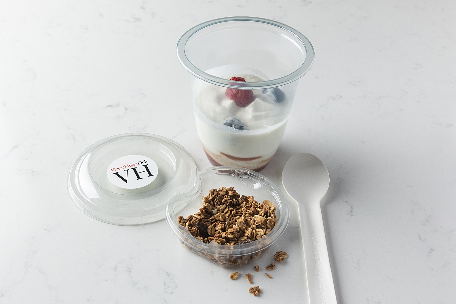 Takeaway yoghurt pot on white marble with granola, fruit and takeaway spoon